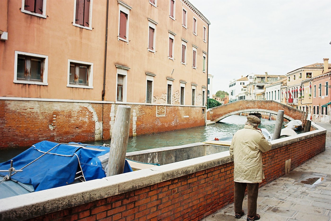 Anziano con berretto e impermeabile chiaro appoggiato al parapetto di mattoni, lo sguardo verso un canale veneziano e un ponte ad arco, barca coperta da un telo blu in primo piano. Fotografia di strada su pellicola a colori 35mm.