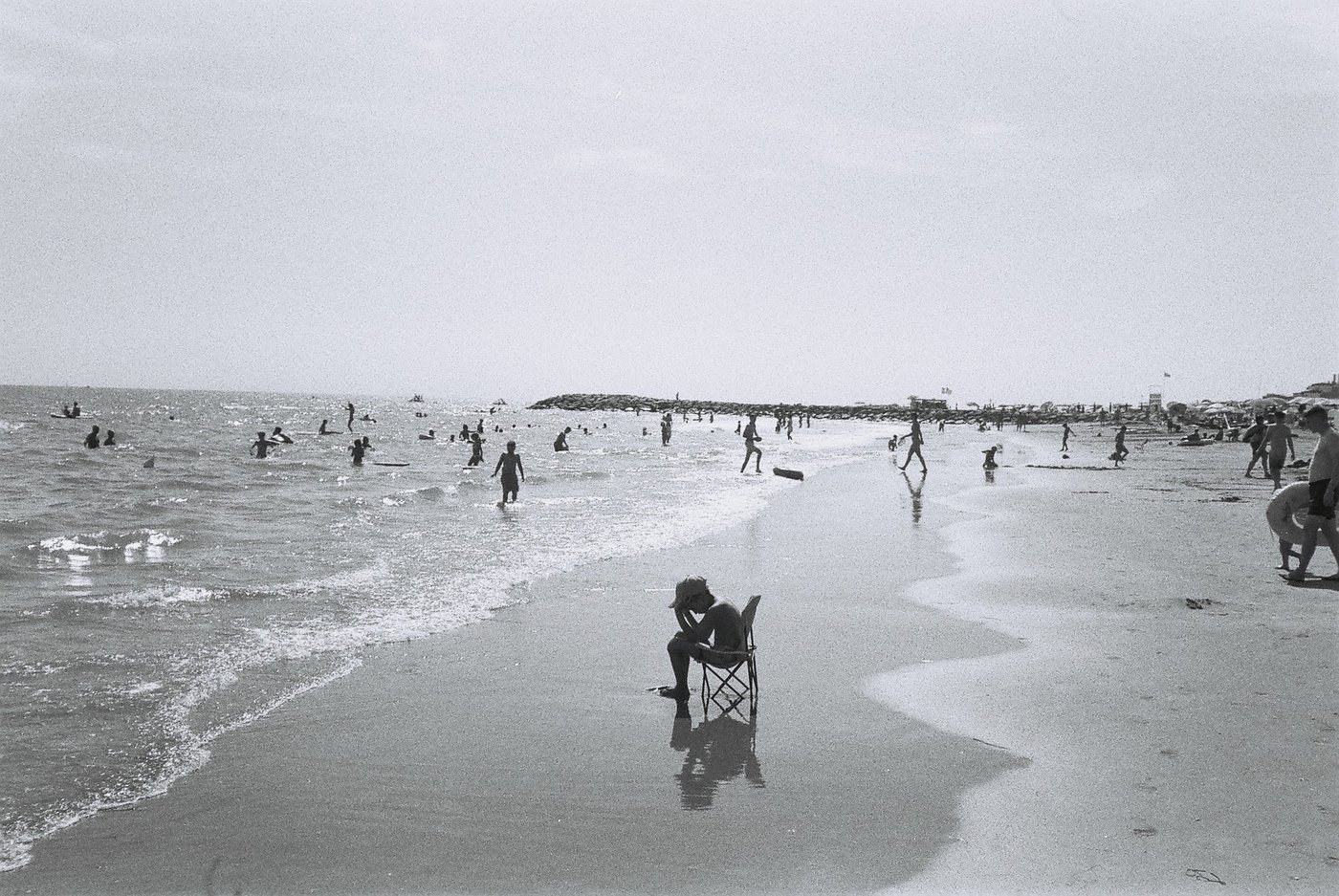 Figura seduta su una sedia pieghevole sulla sabbia bagnata, riflessa sul bagnasciuga, mentre alle spalle bagnanti in controluce entrano in mare verso il frangiflutti. Fotografia analogica in bianco e nero su pellicola 35mm, scattata con Canon Snappy 50.