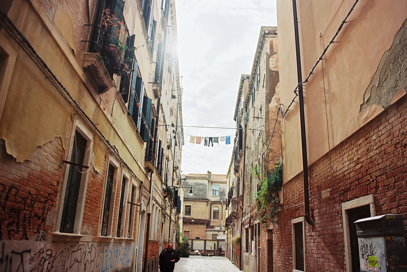 Bucato steso su un filo tra due facciate scrostate di una calle veneziana, intonaco che si stacca dai mattoni e un passante in basso. Fotografia di strada su pellicola a colori 35mm.