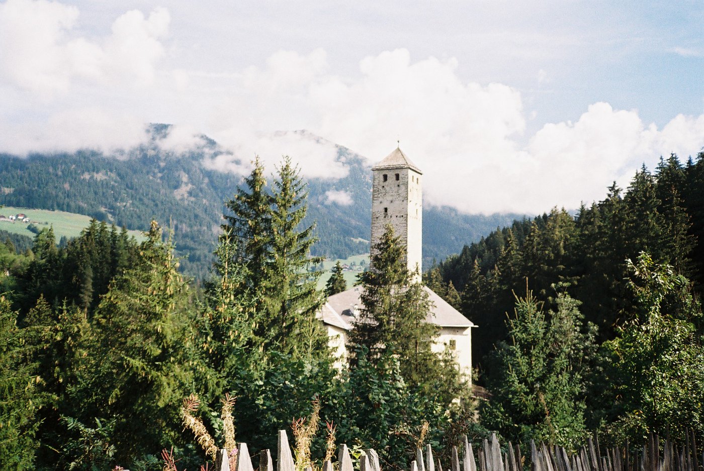 Campanile di pietra che spunta tra gli abeti sotto un cielo carico di nuvole bianche, con le montagne velate dietro. Fotografia analogica di paese alpino su pellicola a colori 35mm.