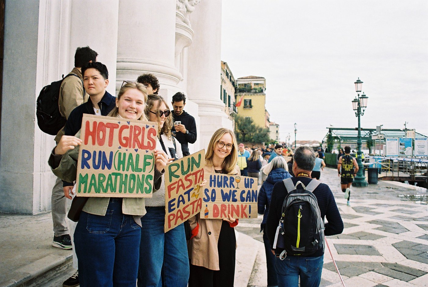 Ragazze sorridenti con cartelli di cartone dipinti a mano che tifano i runner della mezza maratona di Venezia, tra le colonne bianche di una chiesa. Fotografia di strada su pellicola a colori 35mm.
