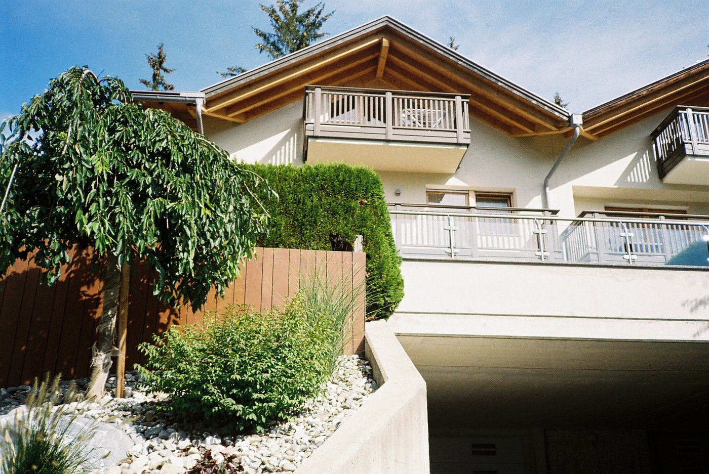 Casa di montagna dalle terrazze in legno chiaro, siepi e ghiaia bianca sotto il sole d'estate. Architettura alpina contemporanea su pellicola a colori 35mm.