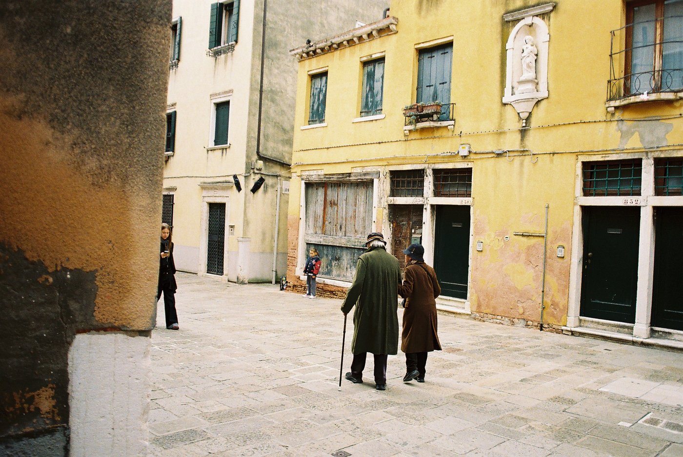 Coppia di anziani che attraversa a braccetto un campo veneziano davanti a una facciata gialla scrostata con una madonnina, lui col bastone; sullo sfondo una bambina e una ragazza col telefono. Fotografia di strada su pellicola a colori 35mm.