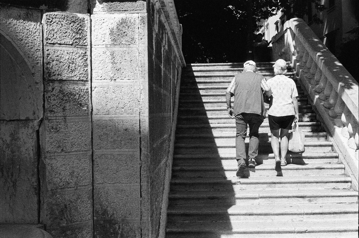 Coppia di anziani che sale a braccetto una scalinata di pietra in pieno sole, tagliata da una lunga diagonale d'ombra accanto a un muro di blocchi bugnati. Fotografia di strada in bianco e nero su pellicola 35mm, scattata con Minolta Hi-Matic 7s.