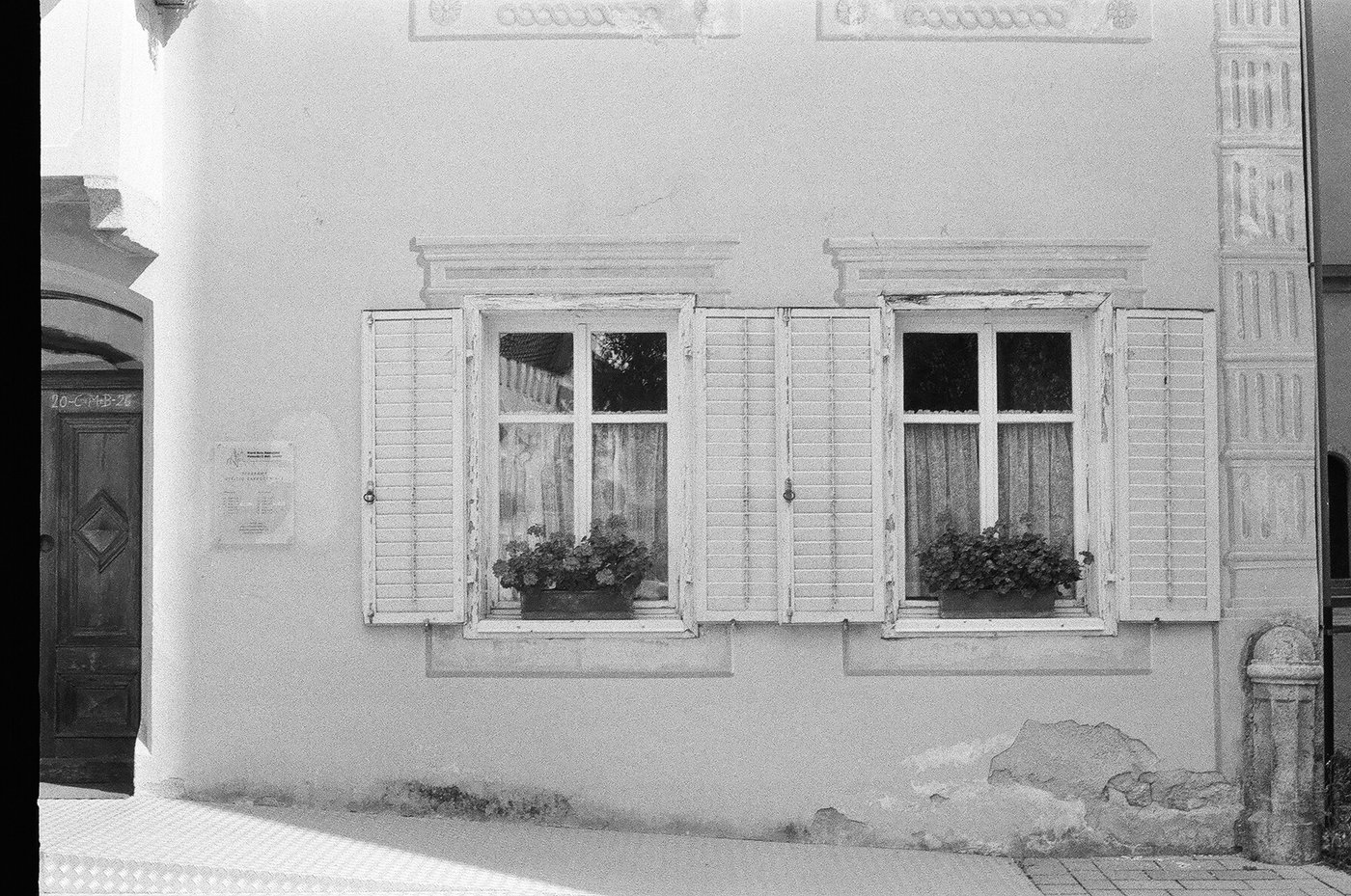 Facciata di casa alpina con due finestre a persiane aperte, vasi di gerani sui davanzali e il portale di legno accanto. Fotografia analogica in bianco e nero su pellicola 35mm, scattata con Minolta Hi-Matic 7s in Alto Adige.