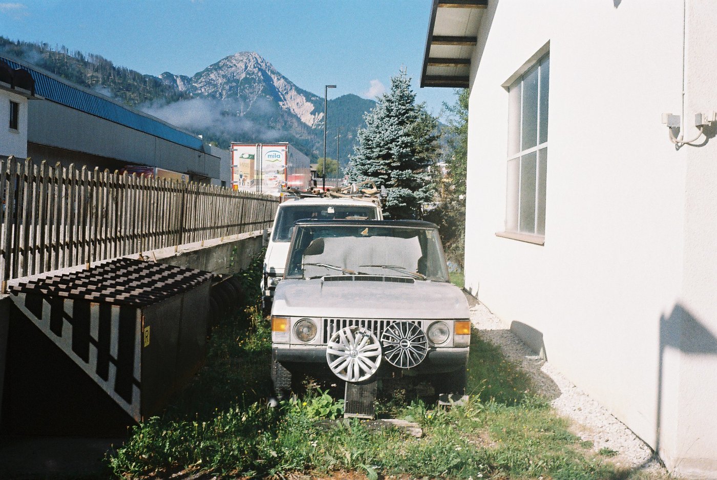 Vecchio fuoristrada bianco parcheggiato tra una staccionata e un muro, con le montagne e le nuvole basse sullo sfondo. Scena quotidiana di paese alpino su pellicola Kodak Gold 200, scattata con Olympus Trip 35.
