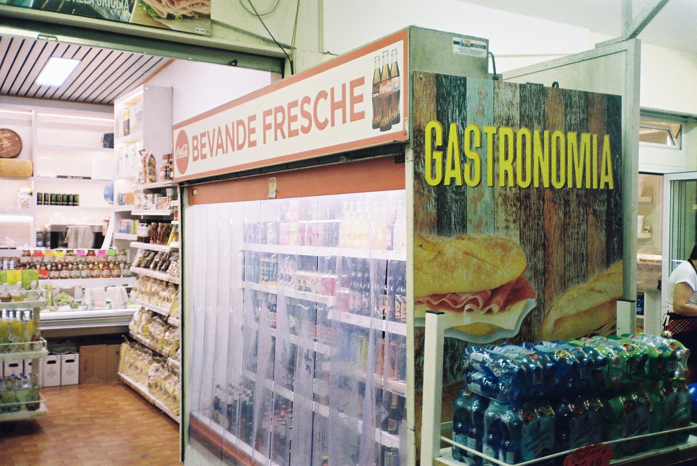 Interno di un minimarket con le insegne vintage Bevande Fresche e Gastronomia, frigo a tendine e casse d'acqua. Scena quotidiana italiana su pellicola 35mm con flash.