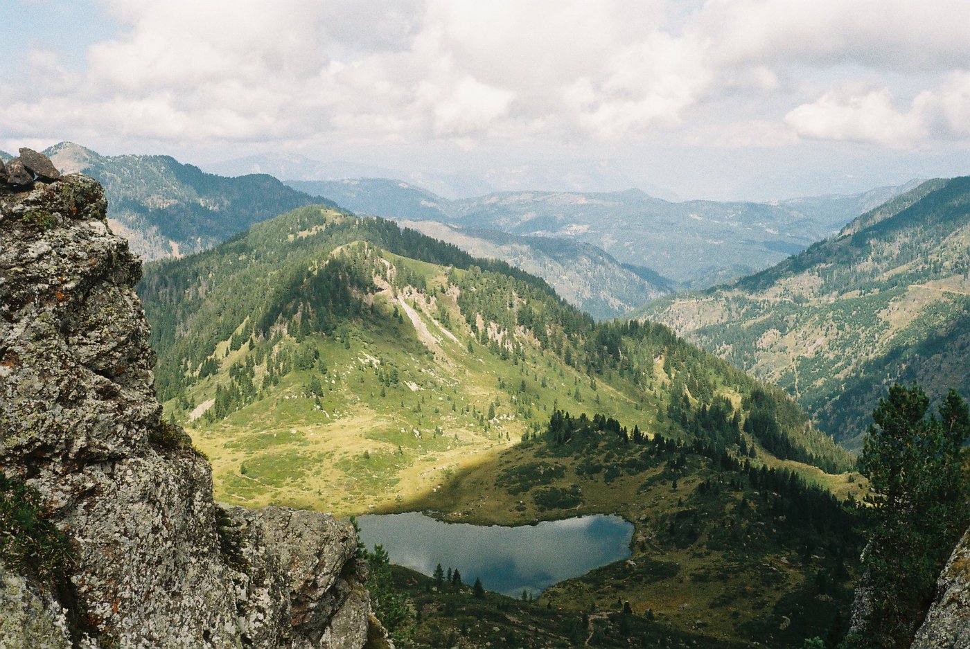 Vista dall'alto su un lago alpino incastonato tra pascoli e boschi, con le valli sullo sfondo. Paesaggio di montagna fotografato su pellicola a colori 35mm.