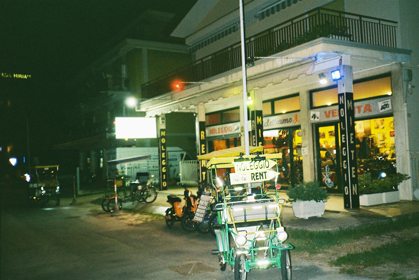 Risciò a pedali davanti a un negozio di noleggio biciclette illuminato nella notte di una località di mare. Fotografia notturna con flash su pellicola a colori 35mm.