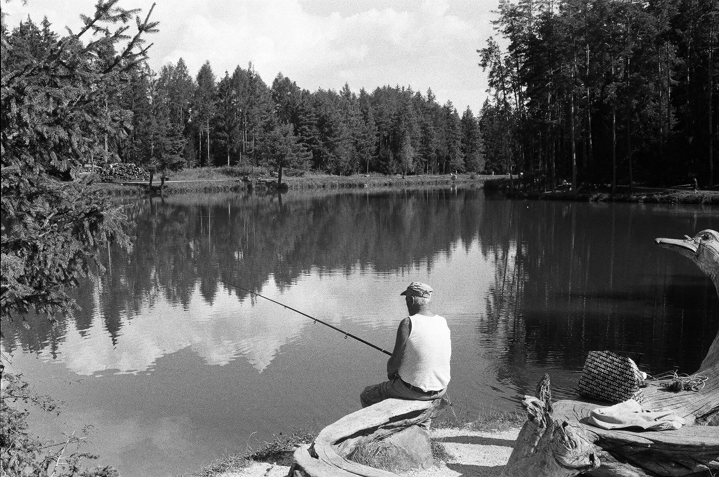 Pescatore seduto su un tronco in riva a un lago alpino, canna tesa sull'acqua ferma che riflette la foresta di abeti. Fotografia analogica in bianco e nero su pellicola 35mm, scattata con Minolta Hi-Matic 7s e obiettivo Rokkor 45mm f/1.8.