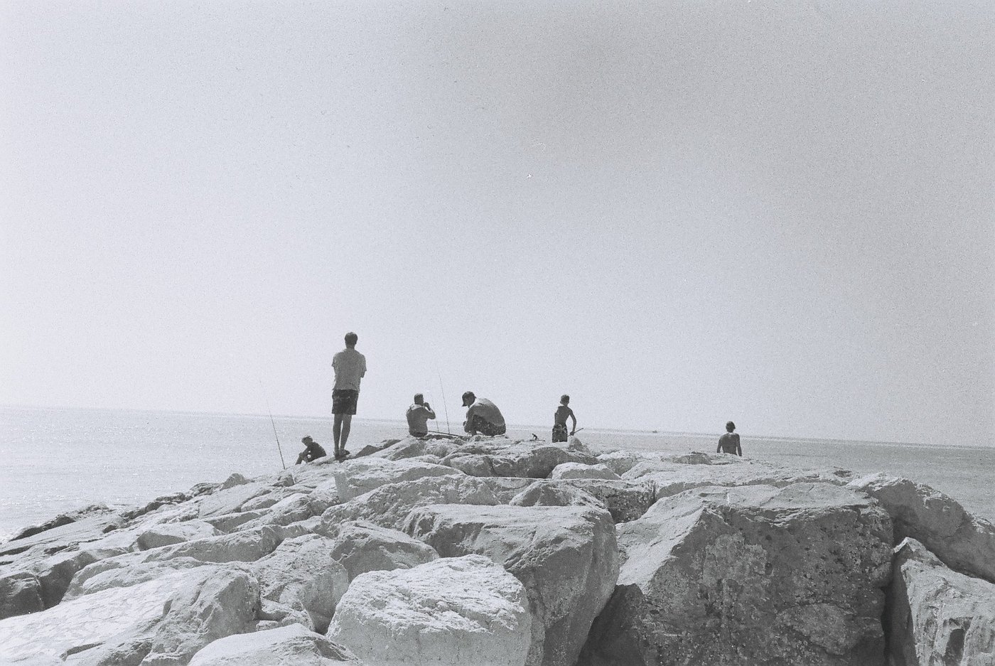 Pescatori e bambini sparsi sui massi di una scogliera, piccole figure contro il mare piatto e un cielo bianco che occupa quasi tutto il fotogramma. Fotografia analogica in bianco e nero su pellicola 35mm, scattata con Canon Snappy 50.