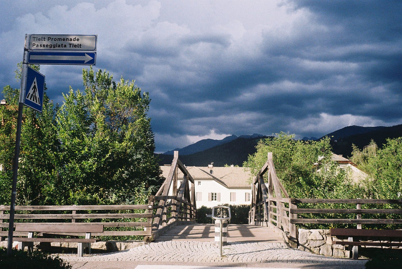 Ponte pedonale di legno illuminato dal sole mentre un cielo nero di temporale copre le montagne alle spalle. Fotografia analogica su pellicola Kodak Gold 200, scattata con Olympus Trip 35.