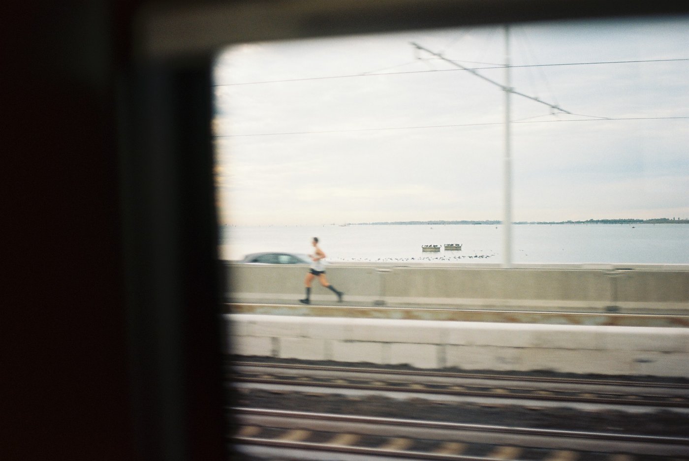 Un runner corre sul Ponte della Libertà visto dal finestrino di un treno in movimento, la laguna di Venezia piatta e grigia sullo sfondo e i binari mossi in primo piano. Fotografia analogica su pellicola a colori 35mm.