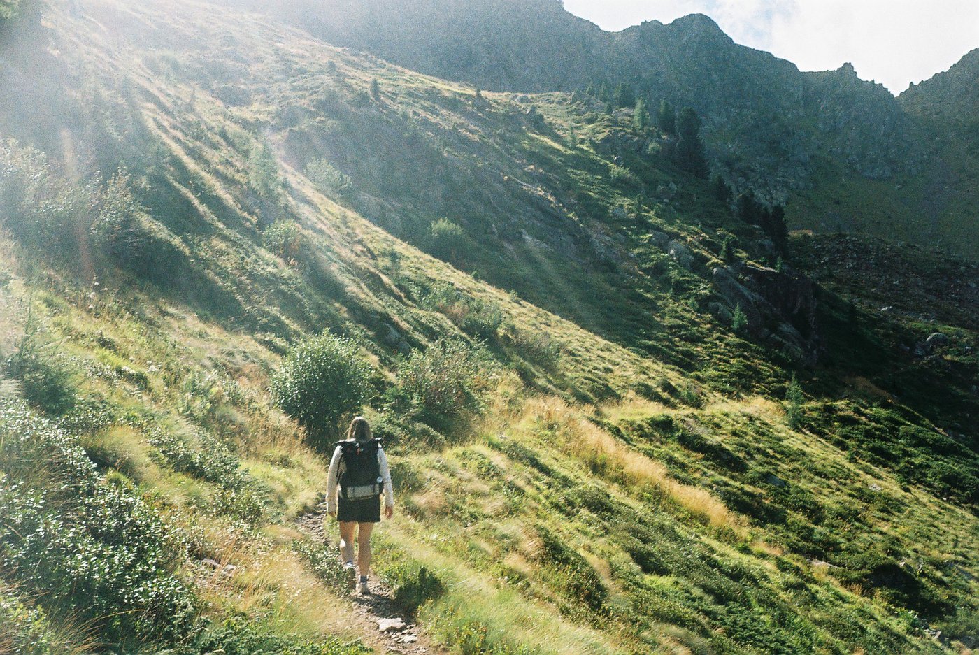 Escursionista in salita su un sentiero alpino, controluce con raggi di sole tra i pendii erbosi. Fotografia analogica su pellicola 35mm a colori.