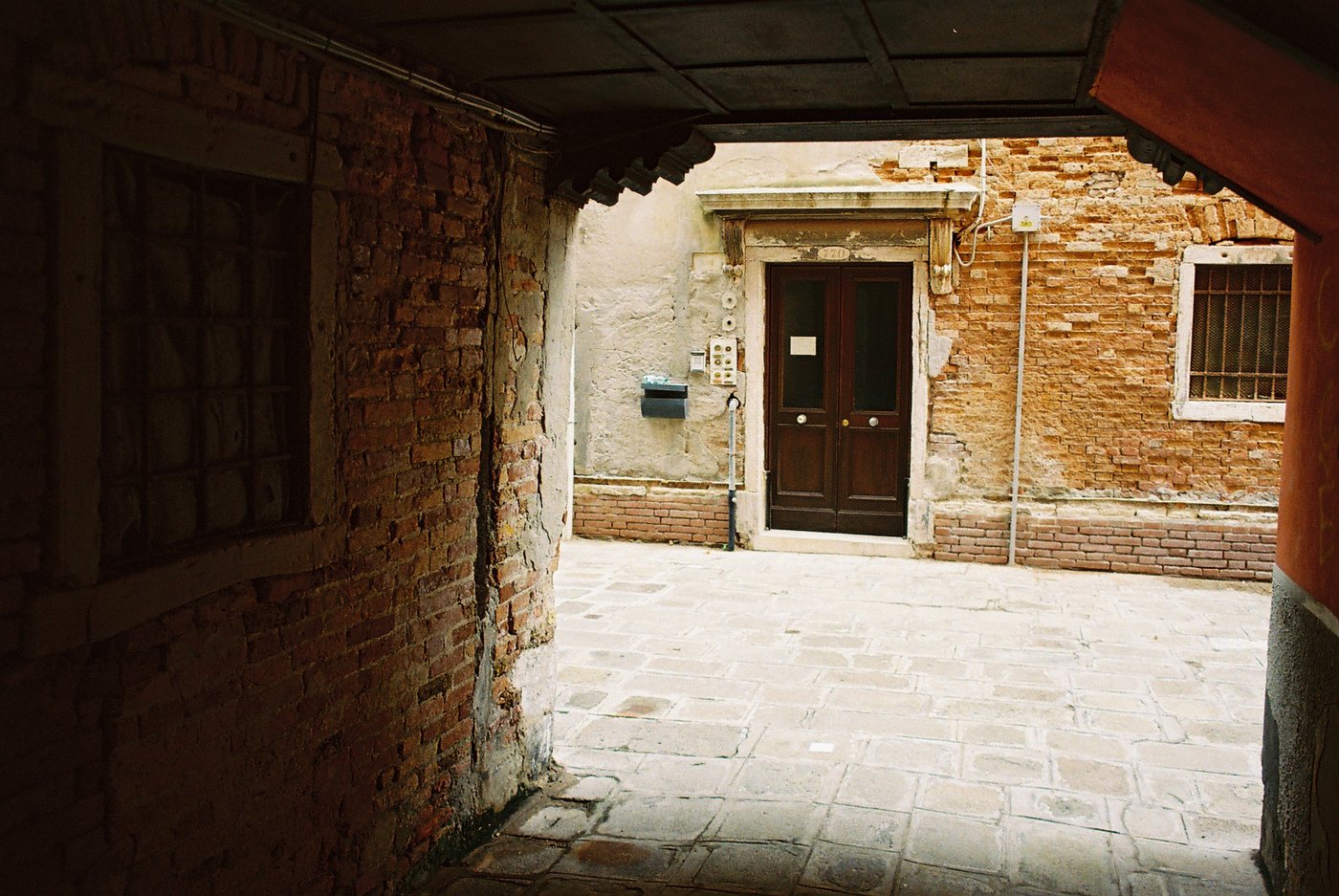 Vista da un sottoportego buio su una piccola corte veneziana illuminata, con una porta di legno scuro incorniciata da pietra d'Istria tra muri di mattoni e intonaco scrostato. Fotografia analogica su pellicola a colori 35mm.