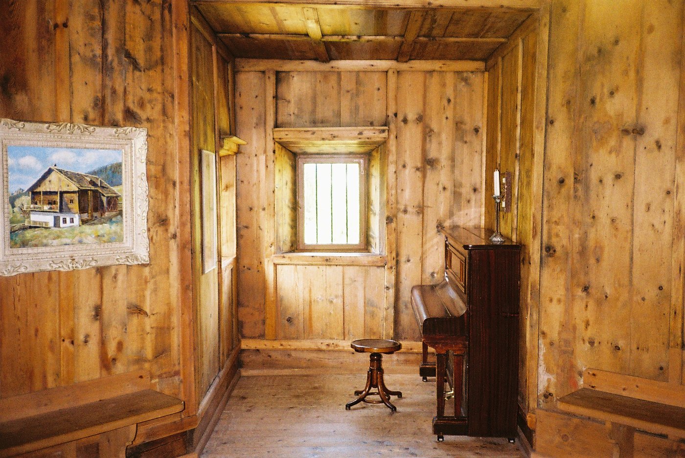 Stube rivestita di legno con un vecchio pianoforte verticale, uno sgabello e una piccola finestra che entra di luce. Interno alpino su pellicola a colori 35mm.