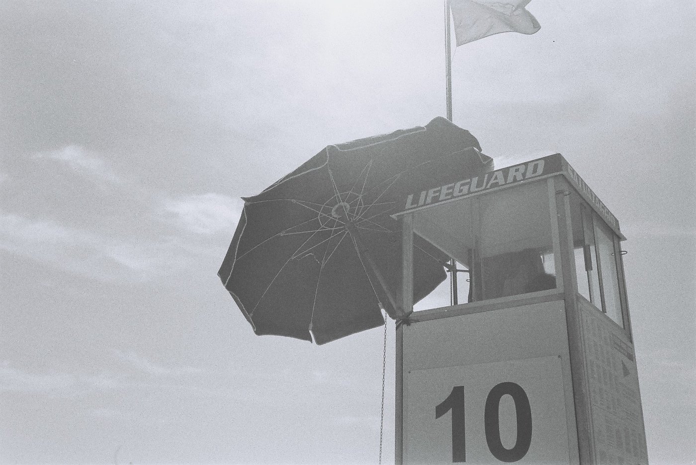 Torretta del bagnino ripresa dal basso, con il numero 10, l'ombrellone aperto e la bandiera mossa dal vento contro un cielo velato. Fotografia analogica in bianco e nero su pellicola 35mm, scattata con Canon Snappy 50.