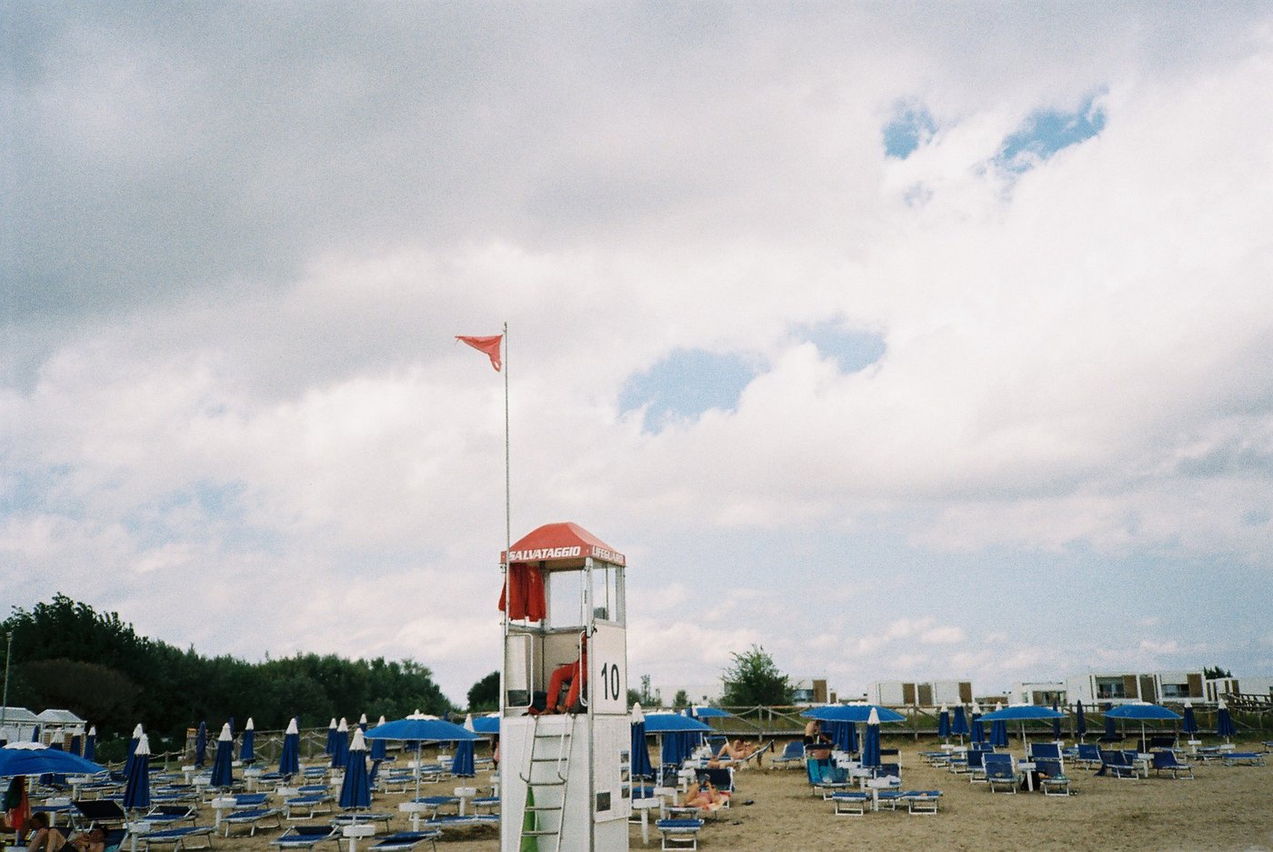 Torretta di salvataggio numero 10 con bandiera rossa al vento su una spiaggia di ombrelloni blu, sotto un cielo carico di nuvole. Fotografia analogica di spiaggia italiana su pellicola 35mm.