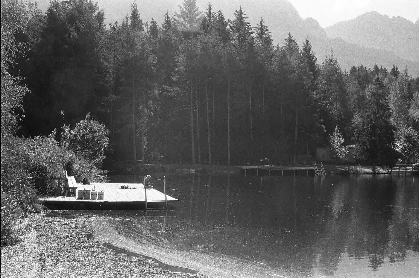 Zattera di legno con una panchina attraccata alla riva di un lago alpino, bosco fitto e cime dolomitiche velate dalla foschia. Fotografia analogica in bianco e nero su pellicola 35mm, scattata con Minolta Hi-Matic 7s in Alto Adige.
