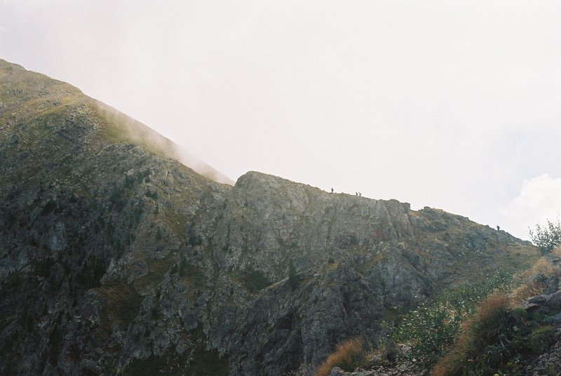 Piccole figure di escursionisti in cammino su una cresta rocciosa mentre la nebbia sale dal versante. Fotografia analogica di montagna su pellicola 35mm.