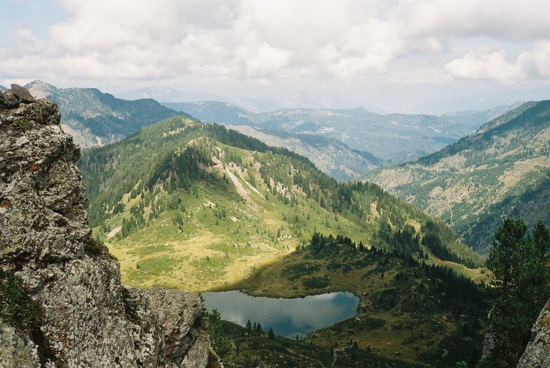 Vista dall'alto su un lago alpino incastonato tra pascoli e boschi, con le valli sullo sfondo. Paesaggio di montagna fotografato su pellicola a colori 35mm.
