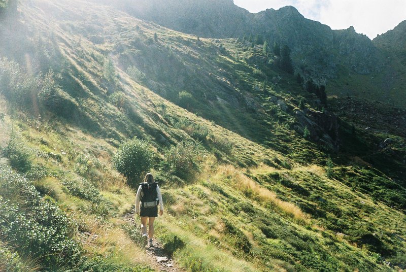 Escursionista in salita su un sentiero alpino, controluce con raggi di sole tra i pendii erbosi. Fotografia analogica su pellicola 35mm a colori.